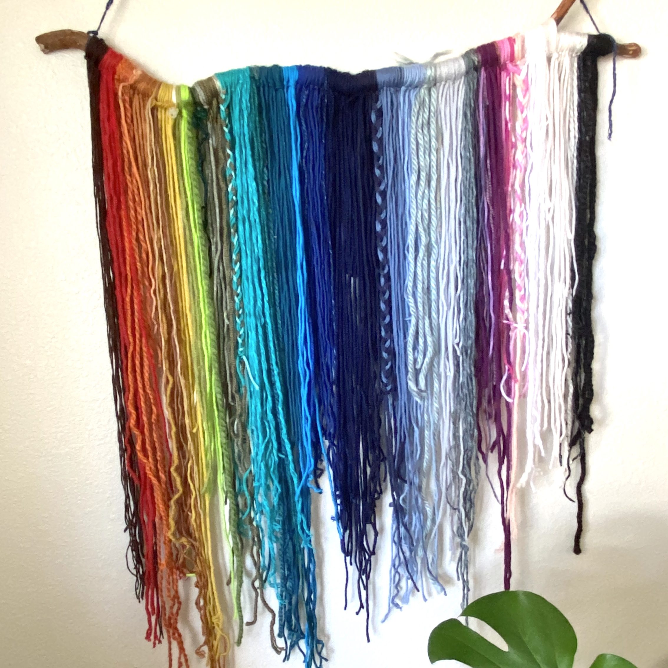 wall-hanging with rainbow yarn hanging from rustic stick with monstera leaf