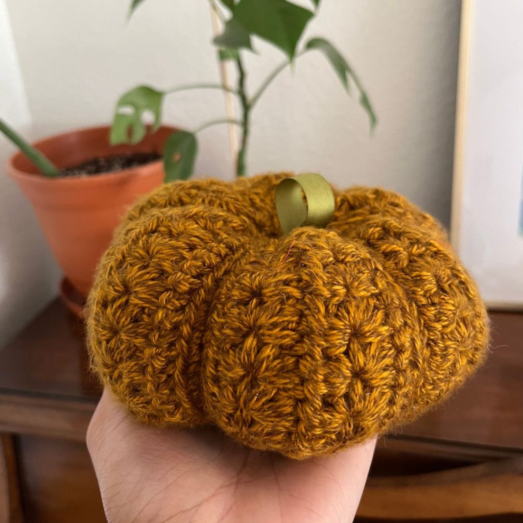 hand holding a brown crochet pumpkin with a green ribbon stem